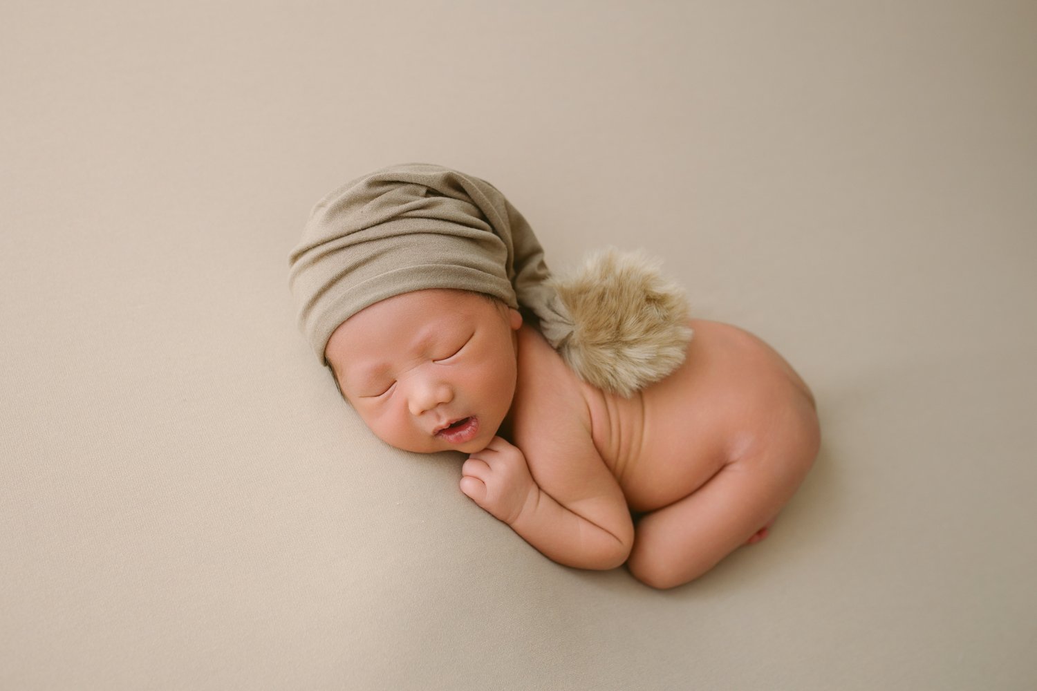 A newborn baby sleeping on a beige surface, wearing a beige hat and a faux fur tail accessory.