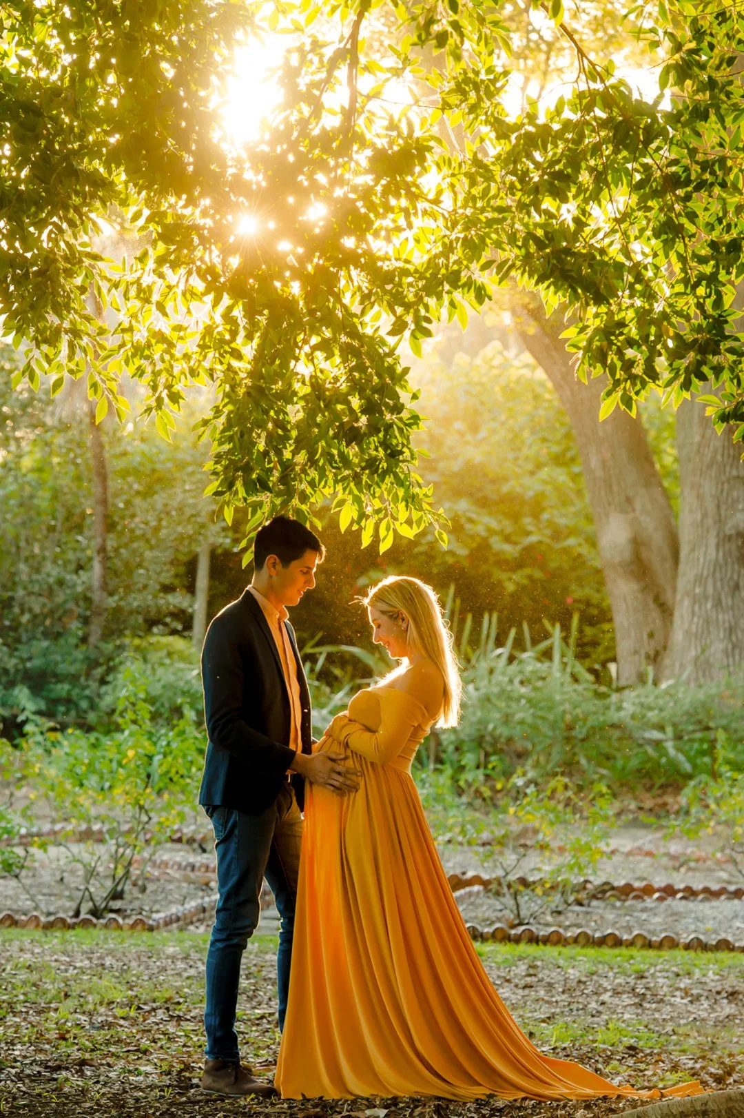 A pregnant woman in a yellow dress standing outdoors with her partner, who is holding her belly, during sunset in a lush, green park.