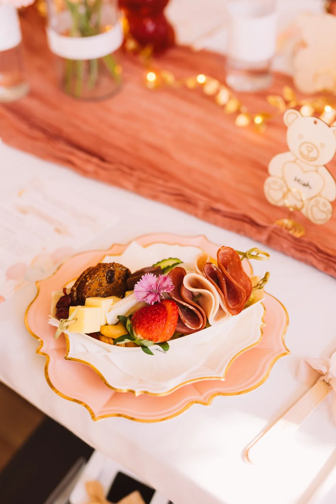 A plate of assorted cheese, strawberries, meats, and crackers on a pink and gold ornate dish placed on a table with a pink tablecloth, with a wooden backdrop and a teddy bear decoration in the background.