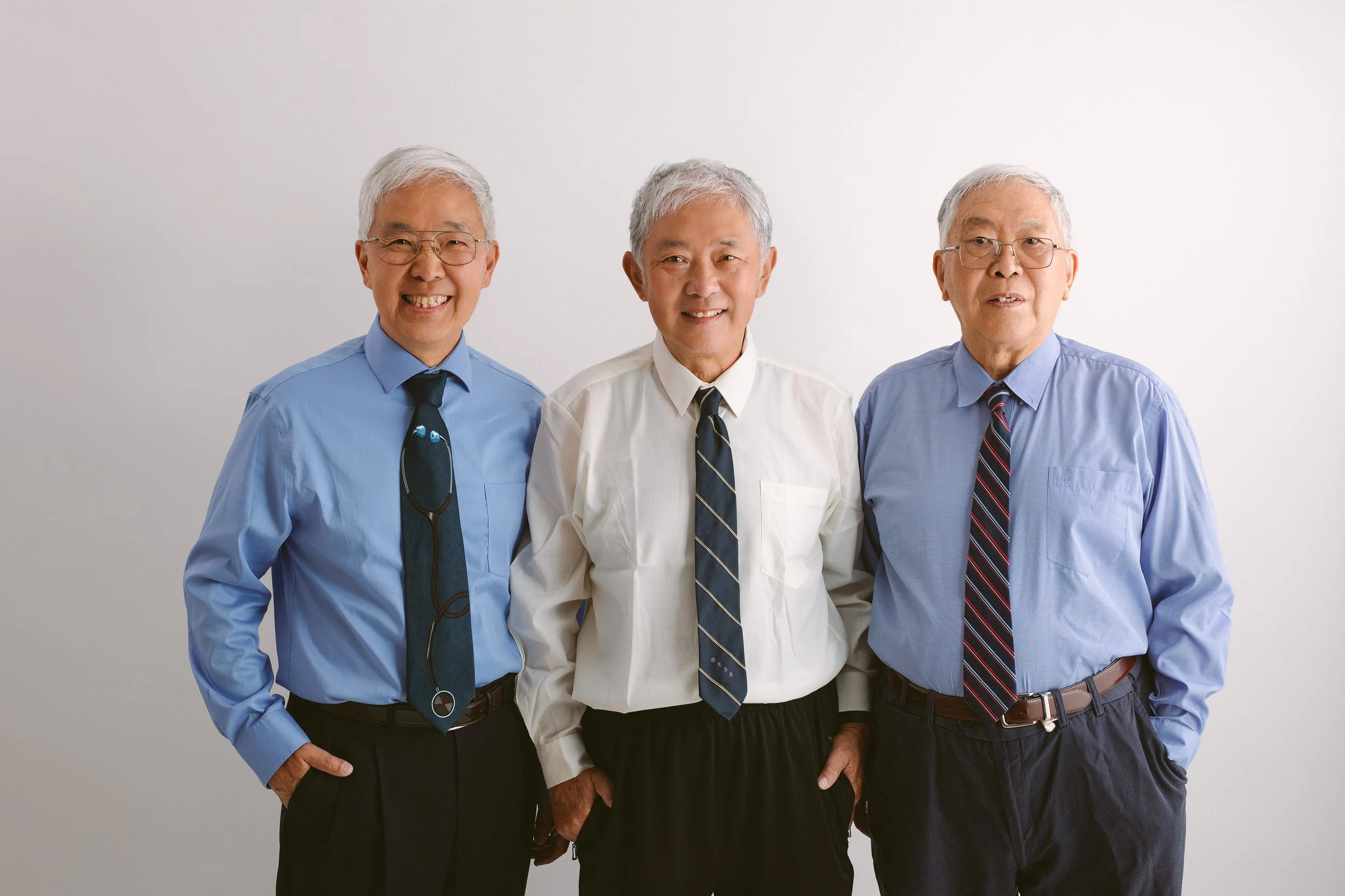 Three elderly men in dress shirts and ties standing together against a plain white background, smiling at the camera.