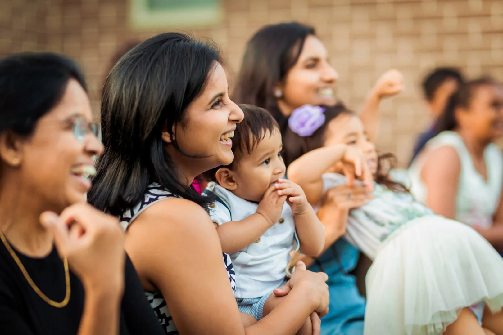Group of women and children watching an event, smiling and seeming entertained, sitting outdoors with a brick wall in the background.