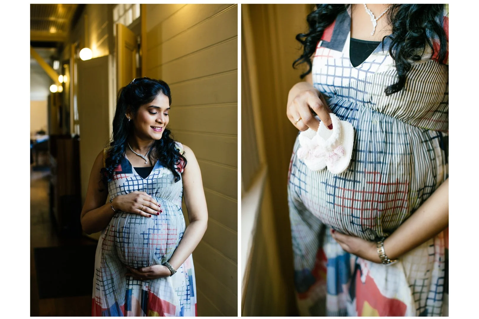 A pregnant woman with long black curly hair in a colorful patterned dress, standing indoors, smiling and holding her belly with one hand and baby shoes in the other.