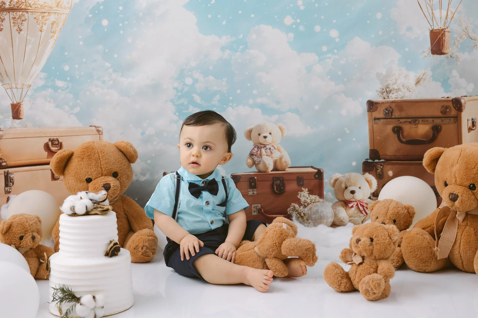 A  one year old baby dressed in a light blue shirt with a black bow tie and suspenders sitting among teddy bears and vintage suitcases, with a sky and cloud backdrop.