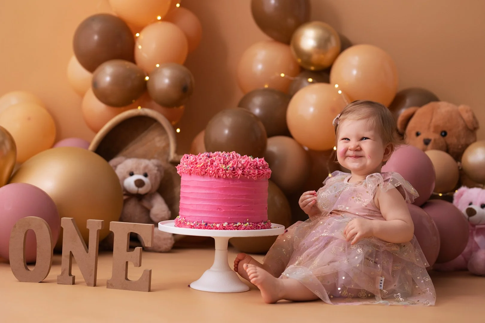 A young girl in a pink dress sitting next to a pink birthday cake with frosting decorations, surrounded by balloons in shades of gold, beige, pink, and brown, teddy bears, and decorative letters spelling "ONE".
