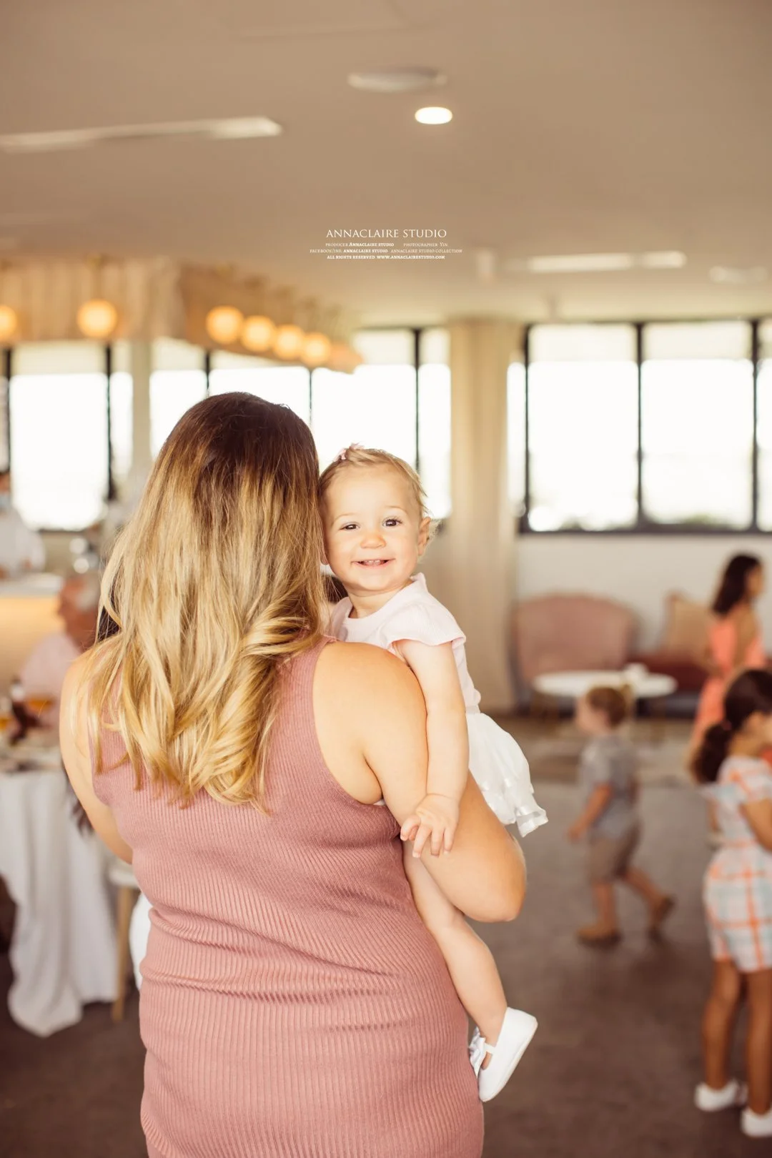 A woman with long, wavy, blonde hair holding a smiling toddler girl in an indoor setting, with other children and adults in the background.