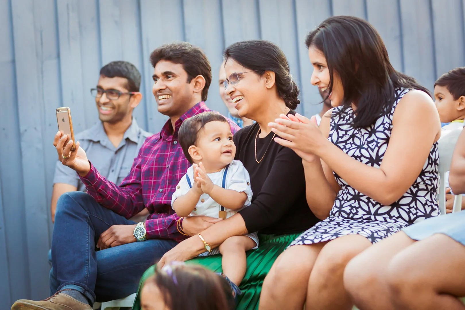 Group of adults and children sitting outside, smiling and enjoying a moment together, with a woman taking a photo or video on her phone.