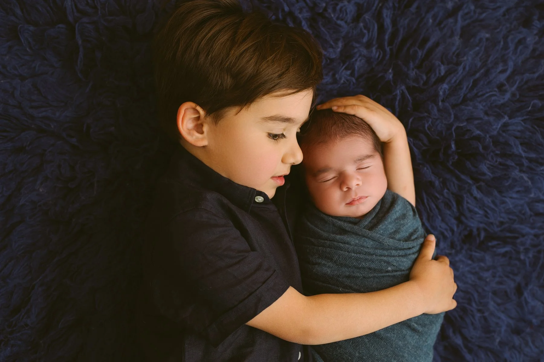 the older brother and one newborn baby, are lying close to each other on a fluffy, dark blue flokati . The older brother is gently holding the baby, and both have peaceful, sleeping expressions.