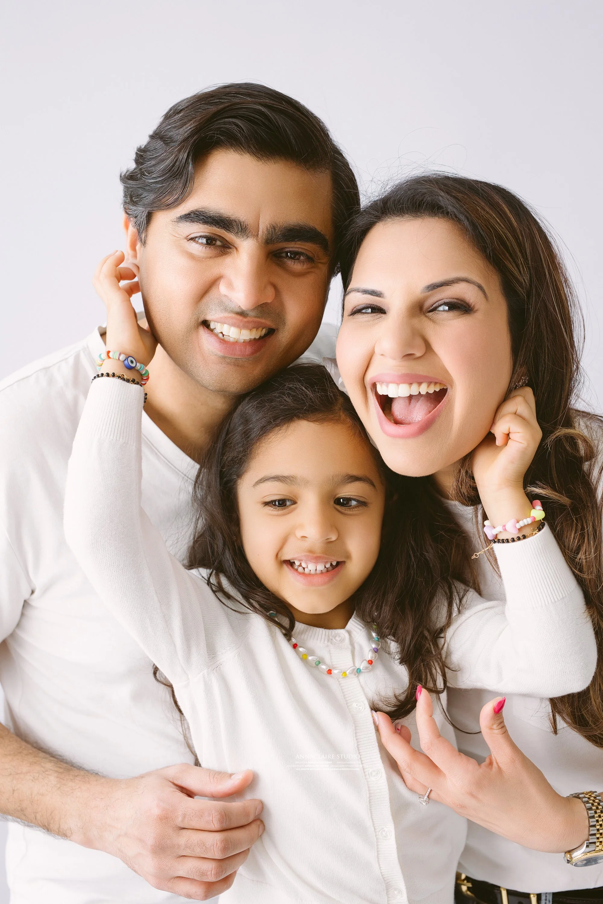 A happy family of three, including a man, woman, and young girl, smiling and embracing each other in a close-up portrait.
