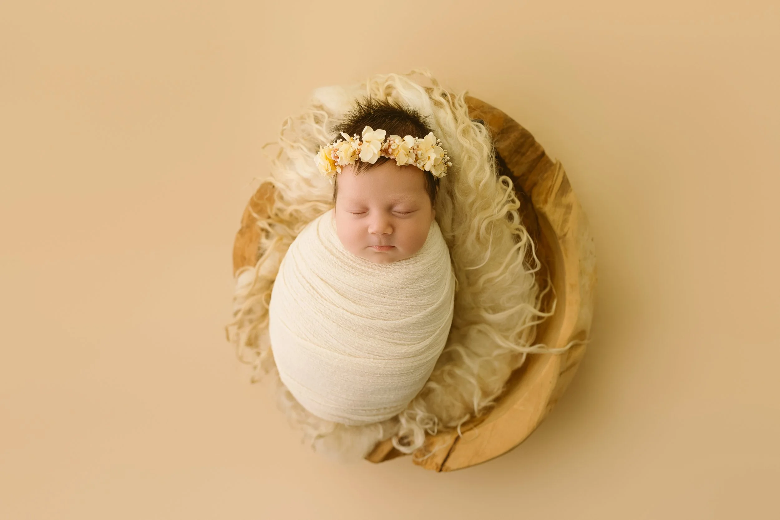 A sleeping baby swaddled in a cream-colored blanket, wearing a flower headband, lying on a round wooden table with fluffy cream-colored fabric surrounding them.