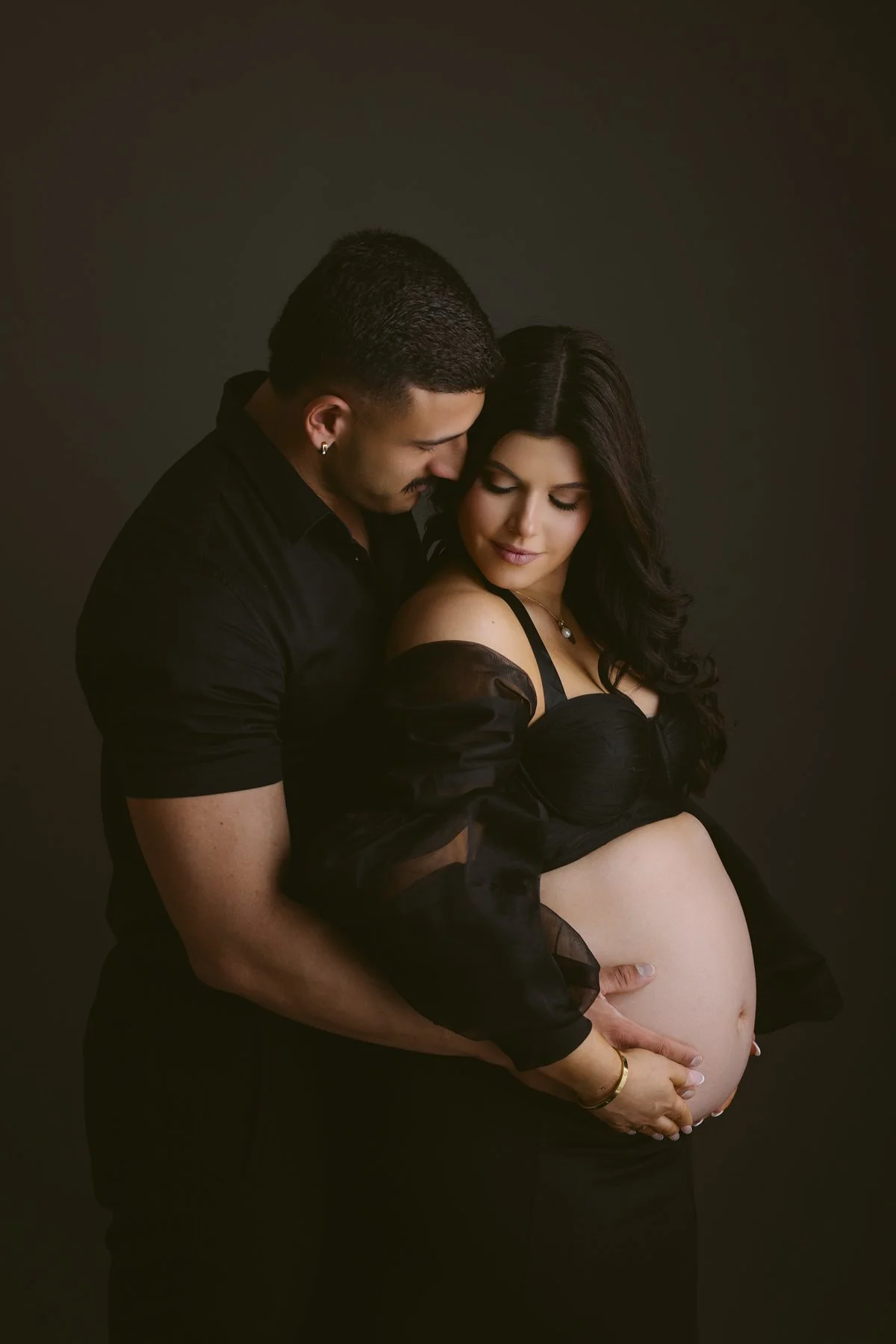 Pregnant woman in black lingerie and sheer robe with a man in black shirt embracing and touching her belly against a dark background.