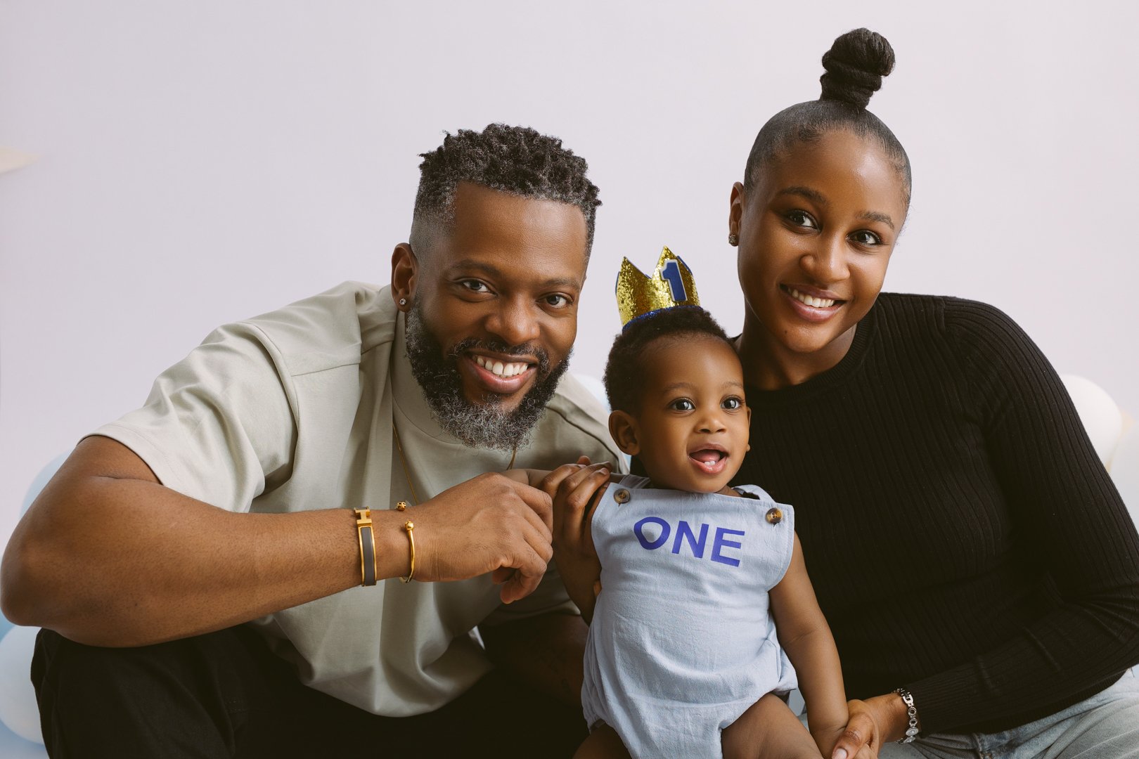 A happy family of three celebrating a child's first birthday, with the child wearing a birthday crown and a shirt with "ONE" on it, posed together indoors.