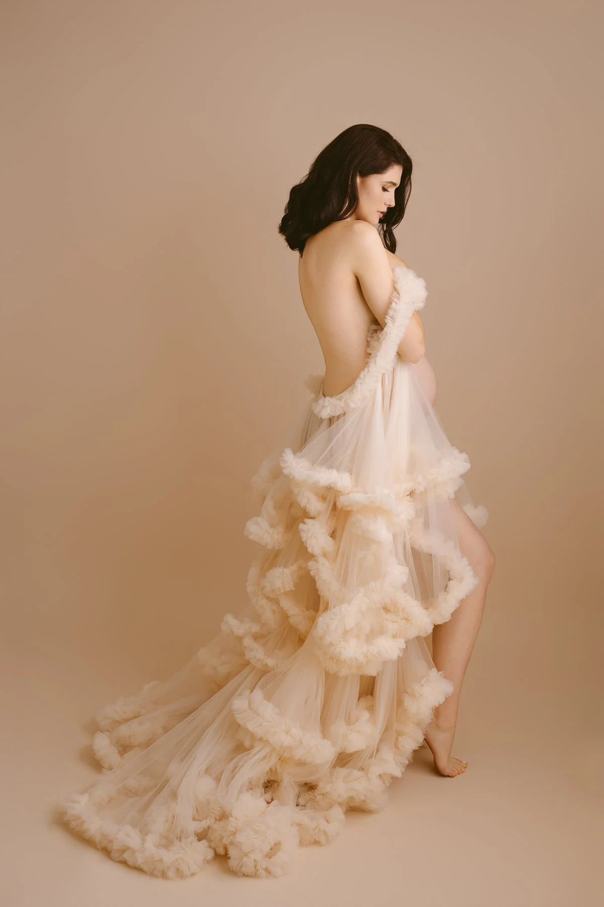 A woman with dark hair standing sideways against a beige background, partially covered with a flowing, cream-colored, fluffy tulle fabric.