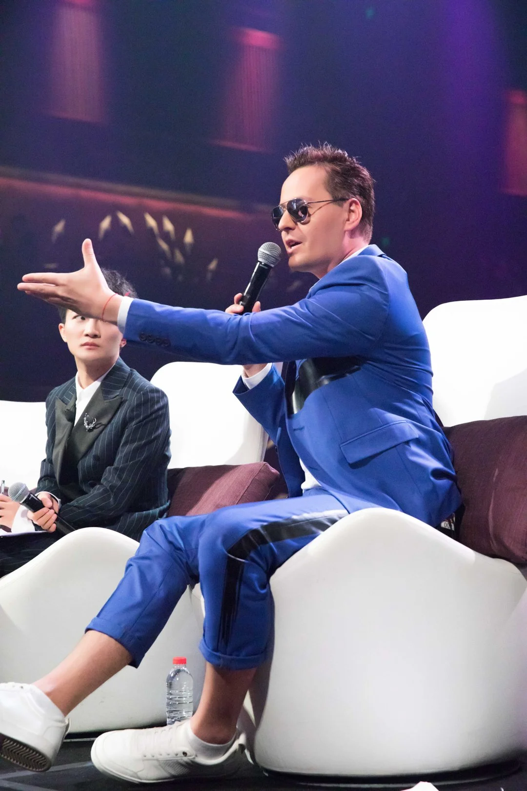 Two men in formal attire participating in a panel discussion, one wearing a blue suit and sunglasses speaking into a microphone, the other in a pinstripe suit holding a microphone, seated on modern white chairs.