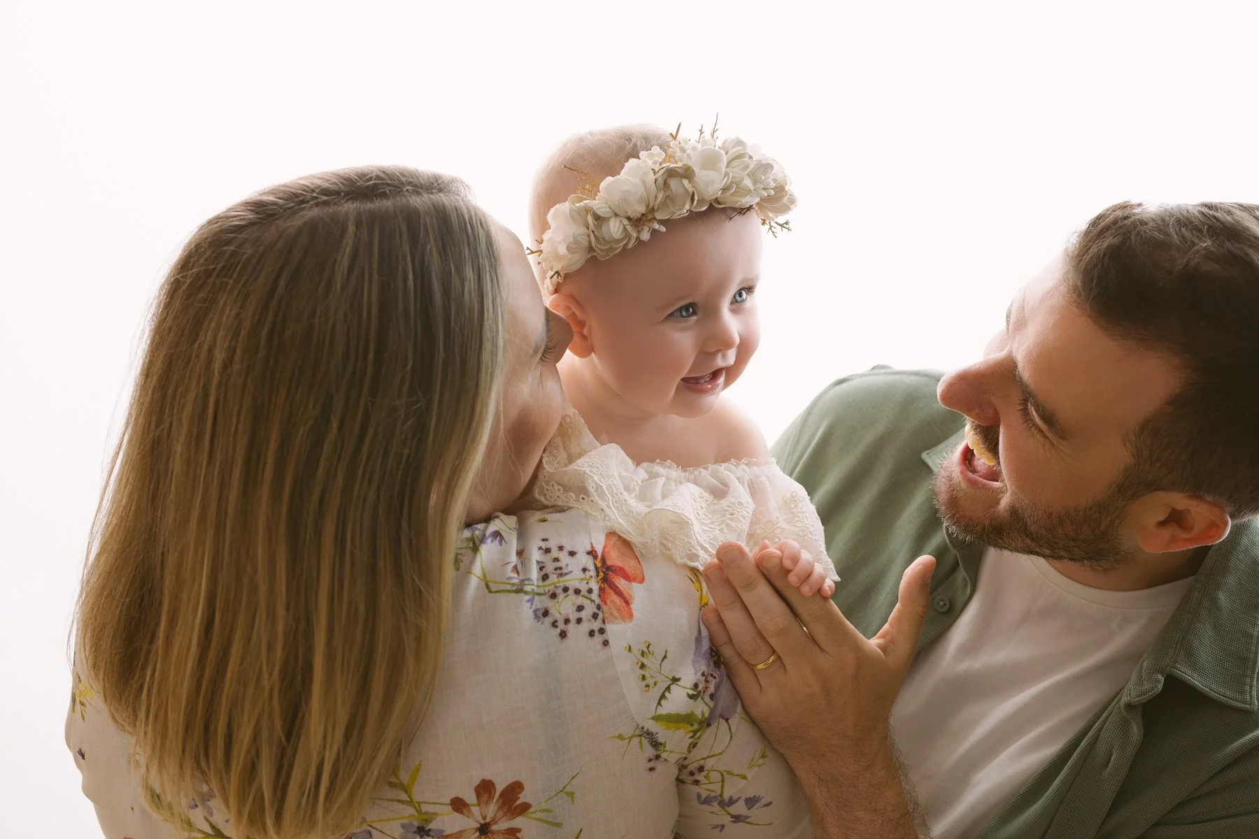 Family of three: mother, father, and a young girl with a flower crown, smiling and looking at each other in a close-up portrait.