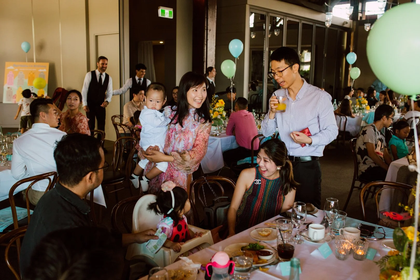 People enjoying a celebration in a decorated indoor venue with tables, balloons, and flowers, including women, men, children, and servers.