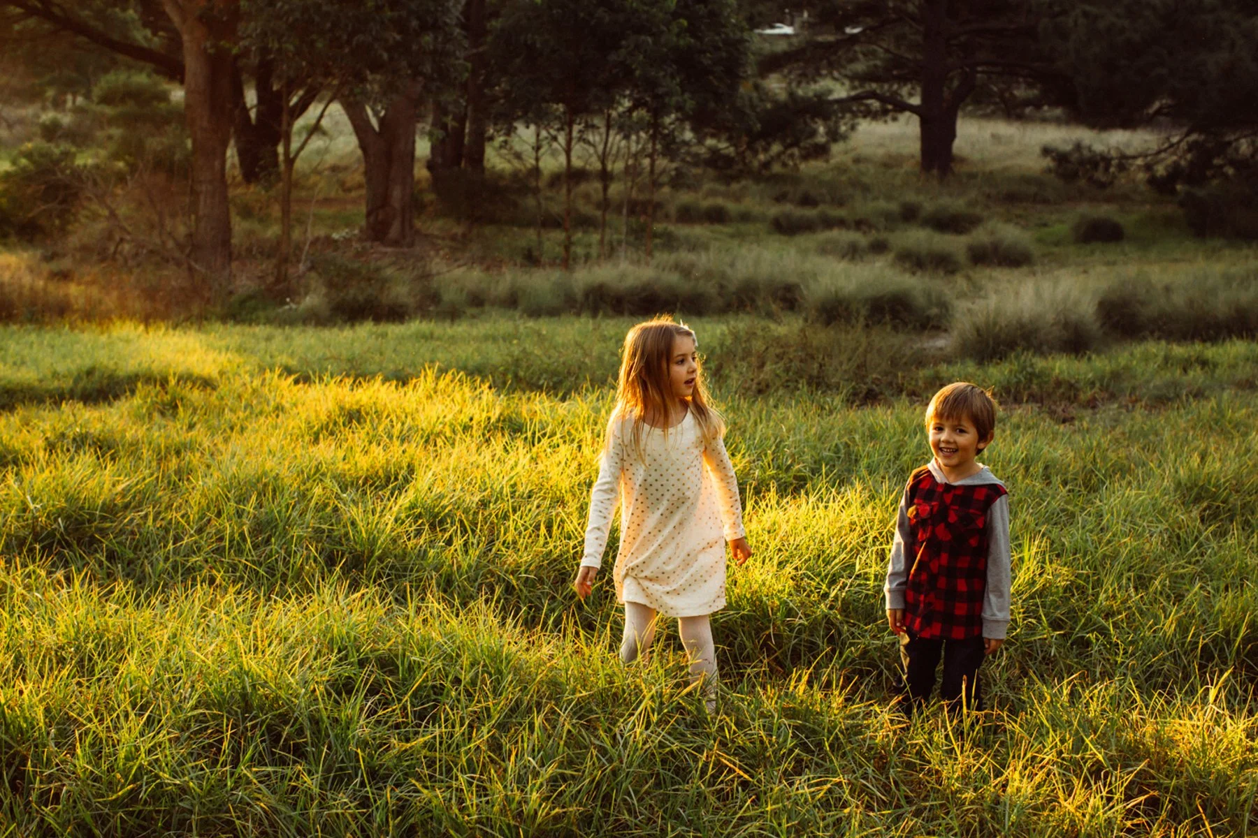 Two young children, a girl and a boy, playing in a grassy field during sunset, with trees in the background.