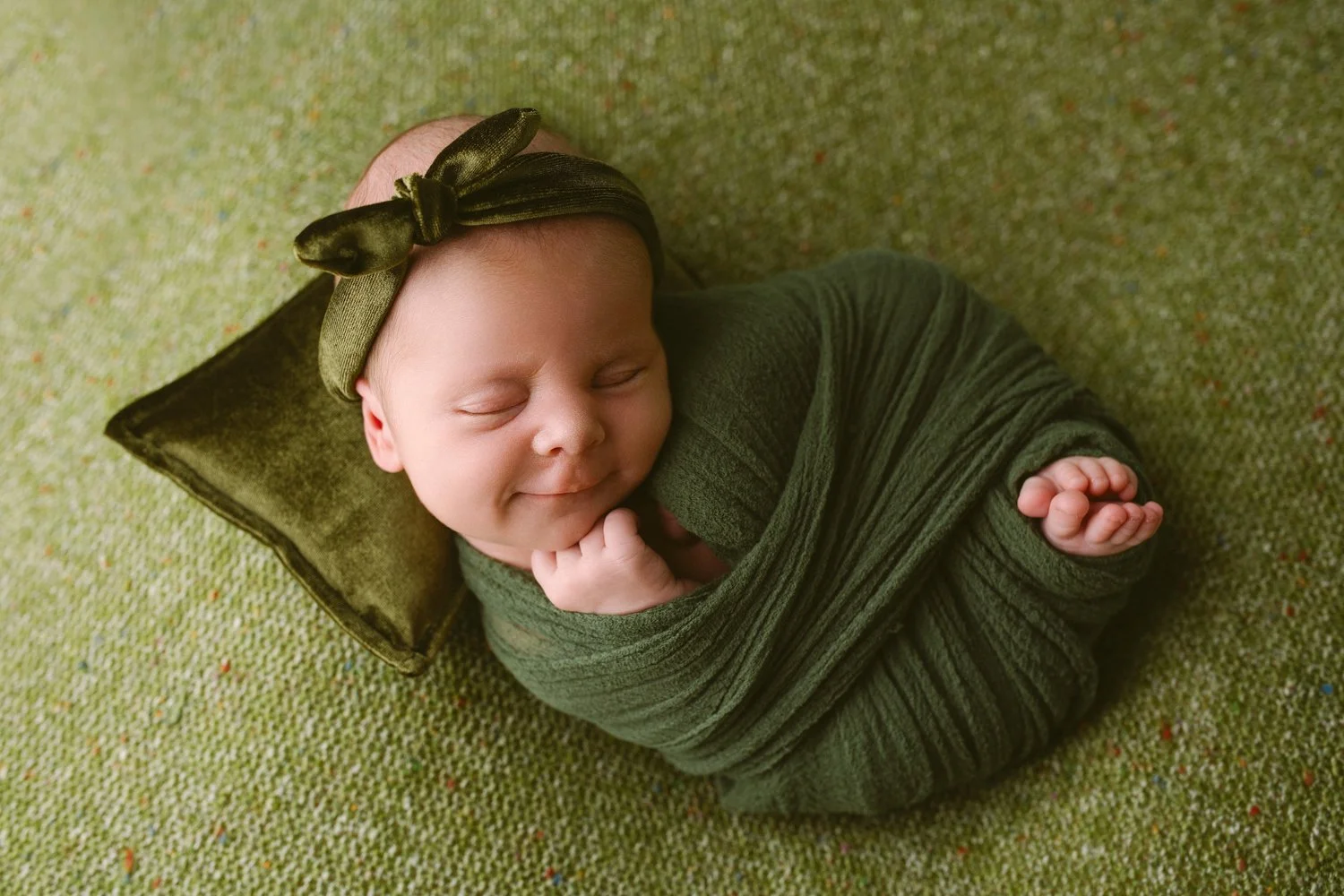 Smiling newborn baby girl  with closed eyes wrapped in green cloth, resting on a small pillow, wearing a matching green headband with a bow, on a green textured surface.
