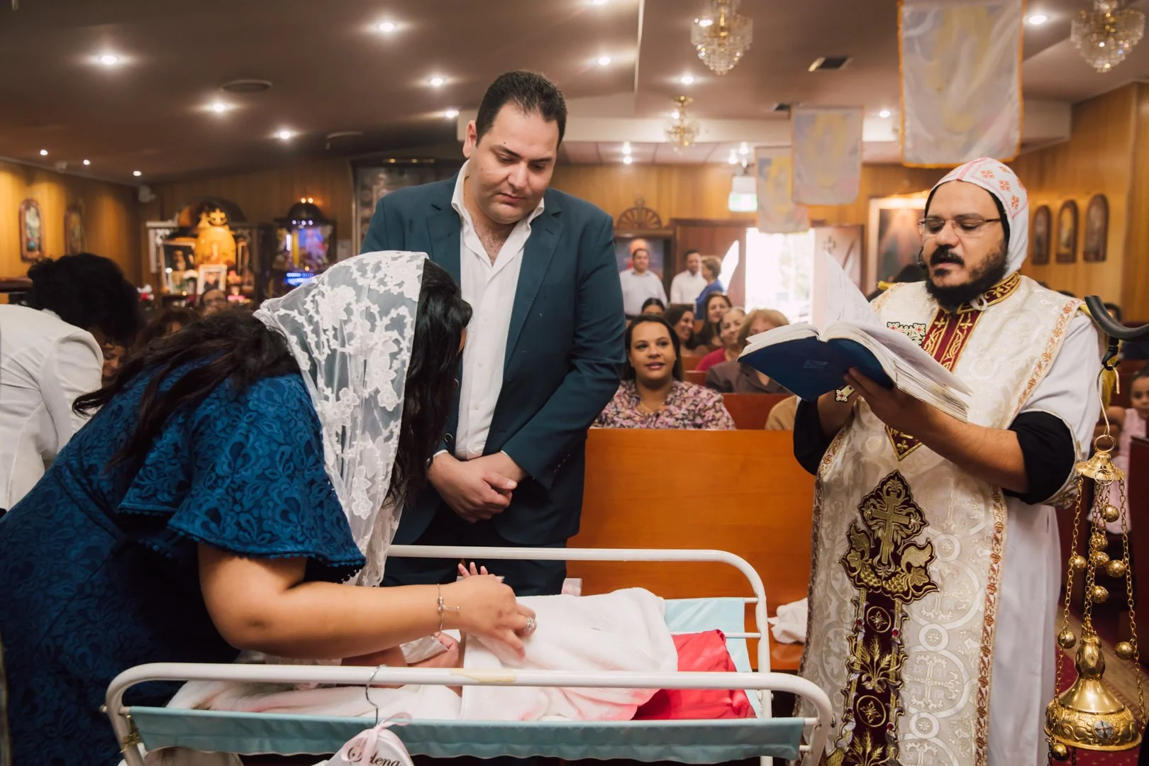 A woman in a blue dress and white lace head covering leans over a hospital crib, holding a newborn baby. A man in a dark suit stands nearby, watching the woman. An Orthodox priest in white and gold ceremonial vestment reads from a book. The scene tak