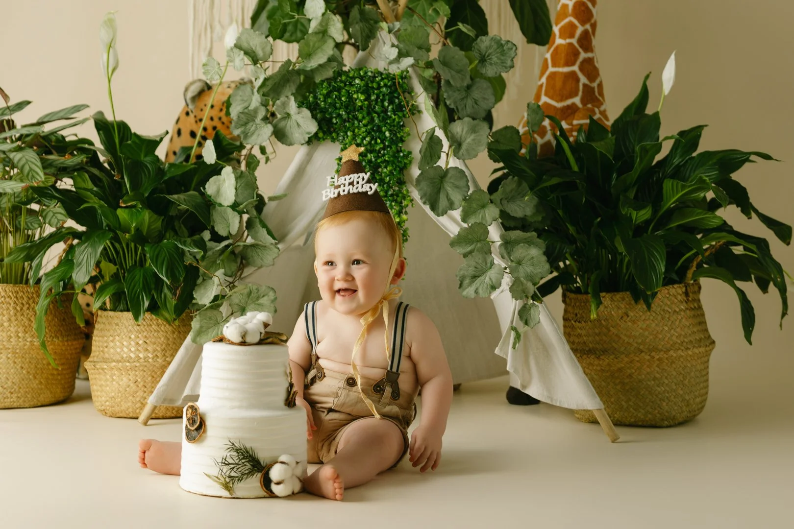 A  one year old baby  sitting on the floor in front of a white birthday cake, wearing a birthday hat with 'Happy Birthday' written on it, surrounded by large green potted plants and a small teepee tent in a decorated indoor setting.