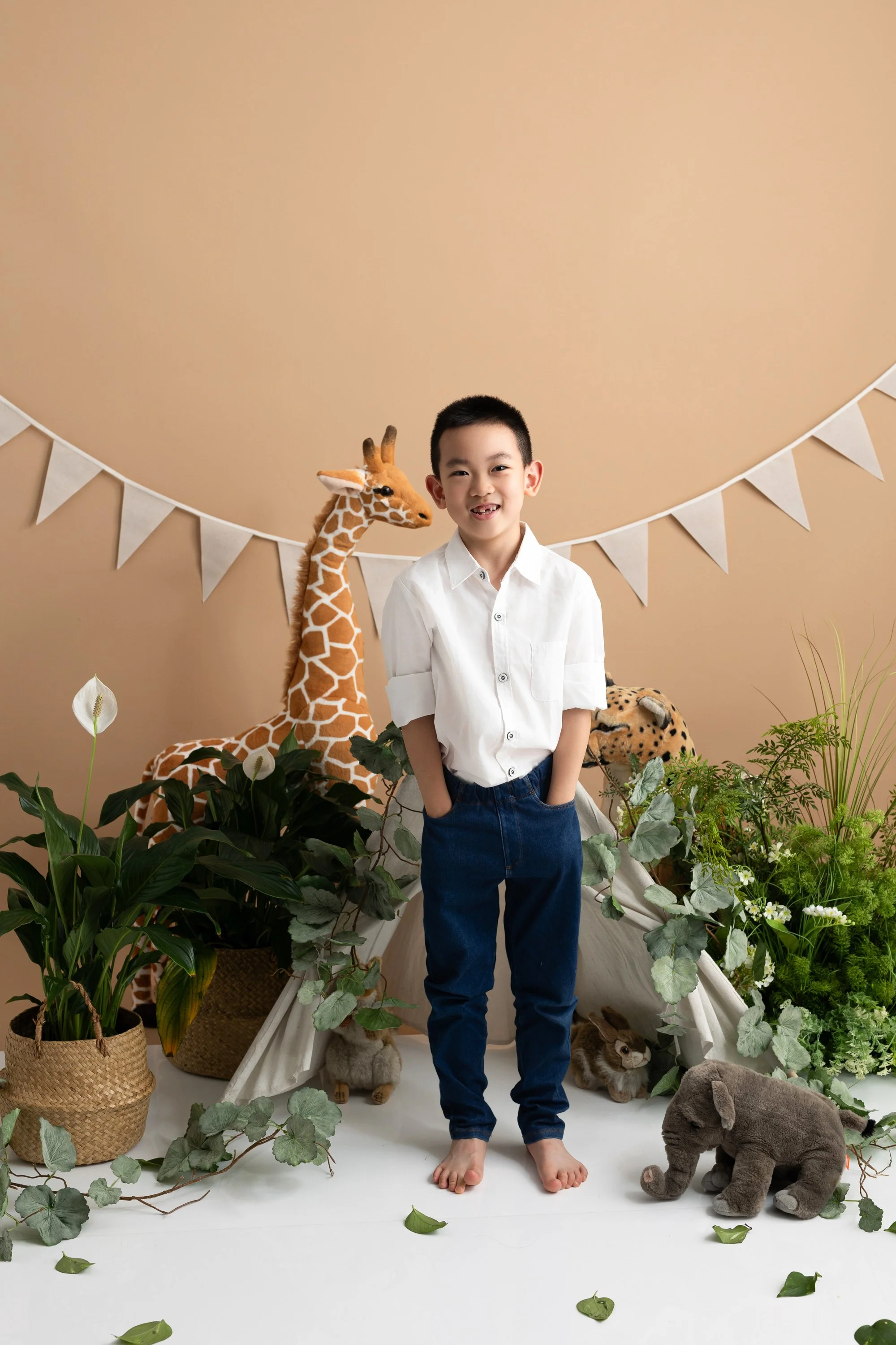 A smiling boy with hands in pockets standing in front of plush animal toys, plants, and a beige background with white bunting.