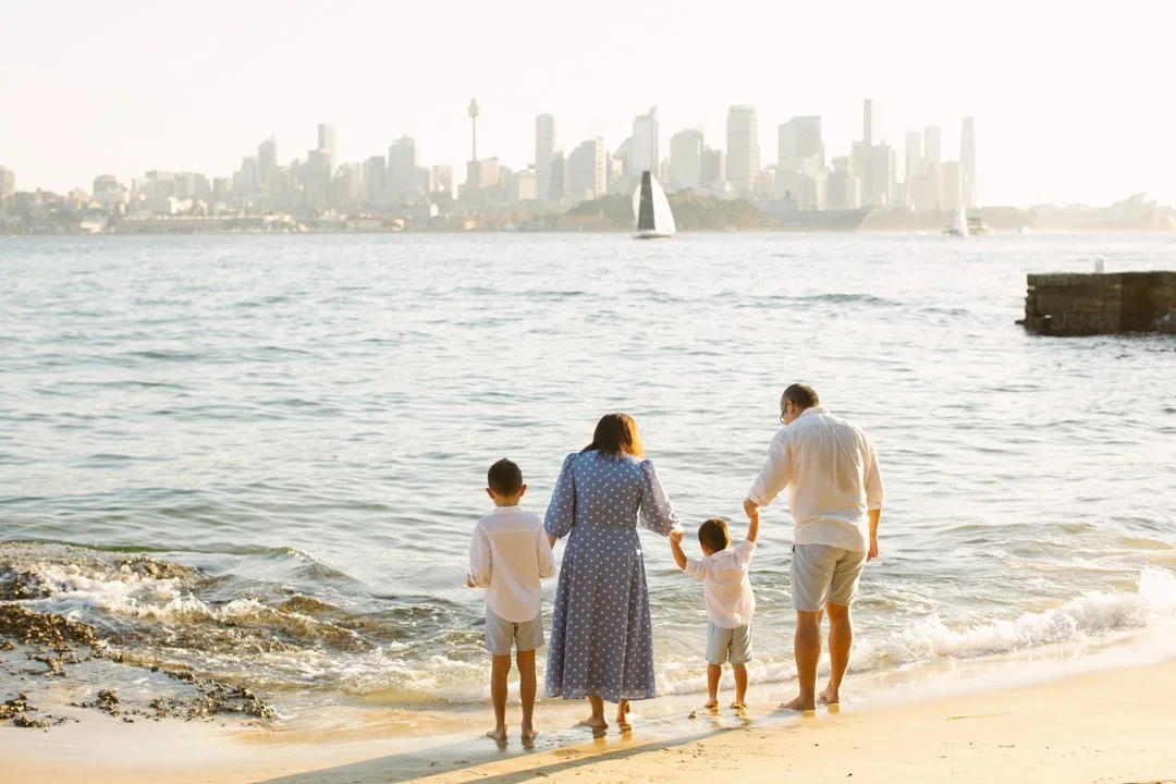 A family of four, including two children, walks hand-in-hand on a beach by the water during sunset with a Sydney city skyline in the background.