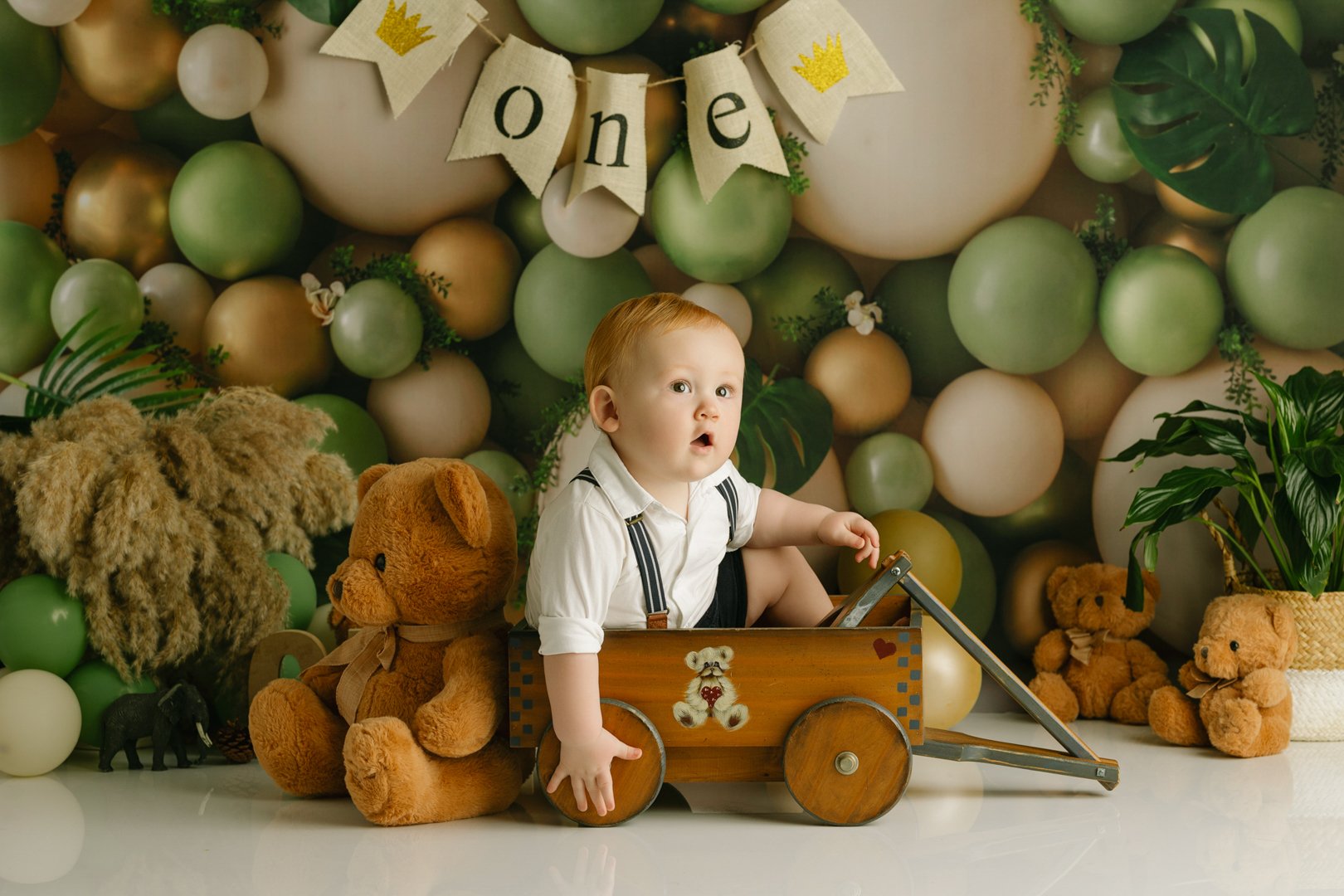 1 yr old  boy sitting in a wooden toy wagon surrounded by teddy bears and plush animals in front of a background decorated with balloons in shades of green, white, and gold, and a banner with the word 'one'.