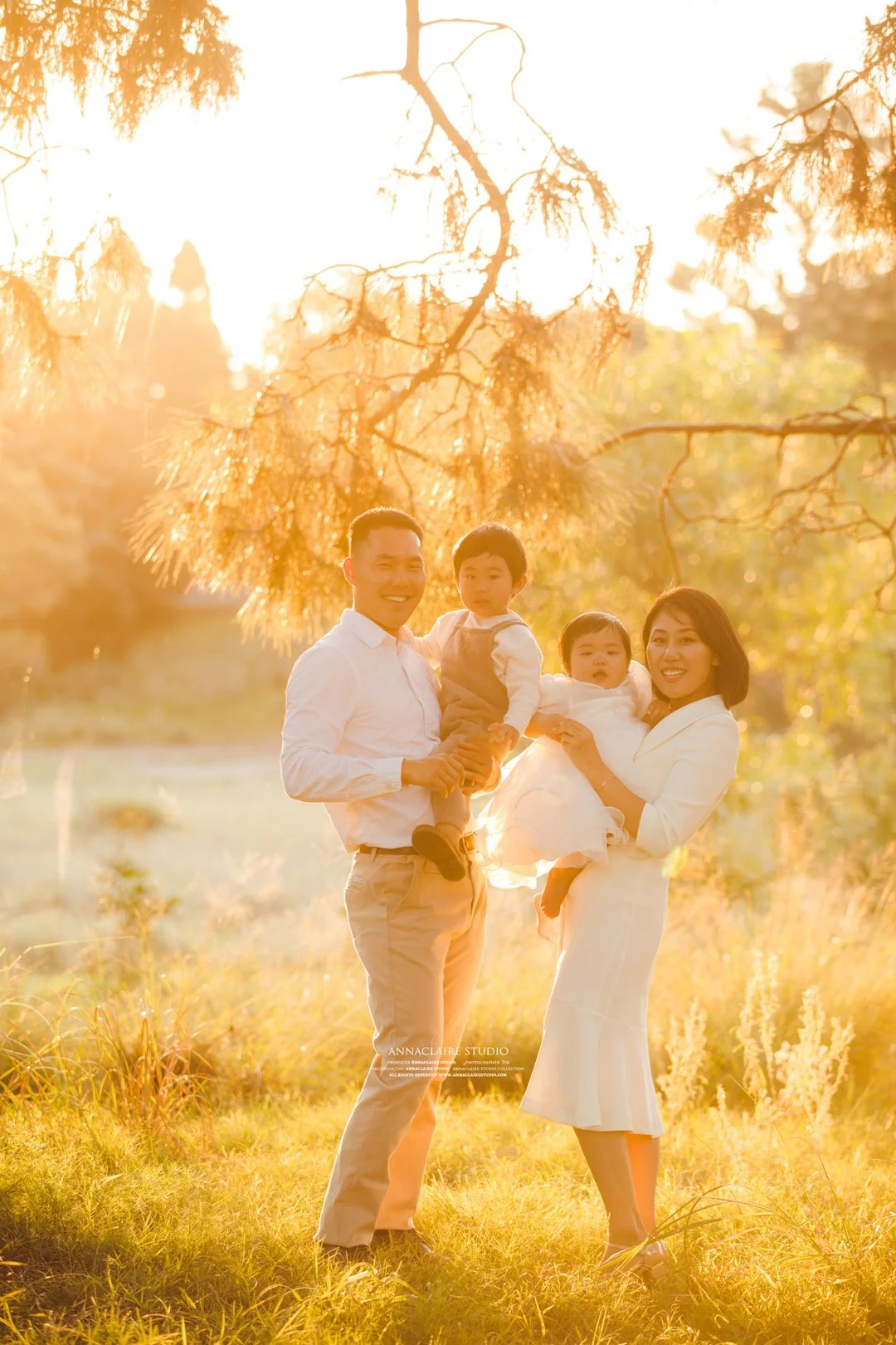 A family of four standing outdoors in a field during golden hour, with trees in the background. The father and mother are holding their two young children, all smiling and dressed in light-colored clothing.