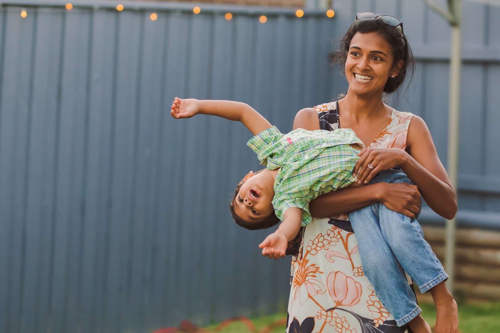 A woman smiles while holding a young boy in her arms outside. The boy appears to be in motion, with his arms outstretched and his mouth open. They are in front of a blue fence and string lights are visible above.