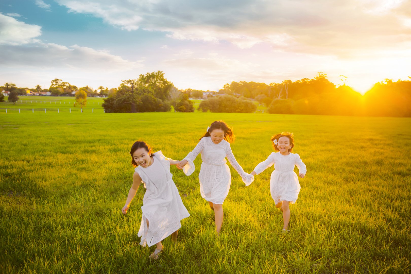 Three young girls wearing white dresses run and play in a green field during sunset, smiling and holding hands.