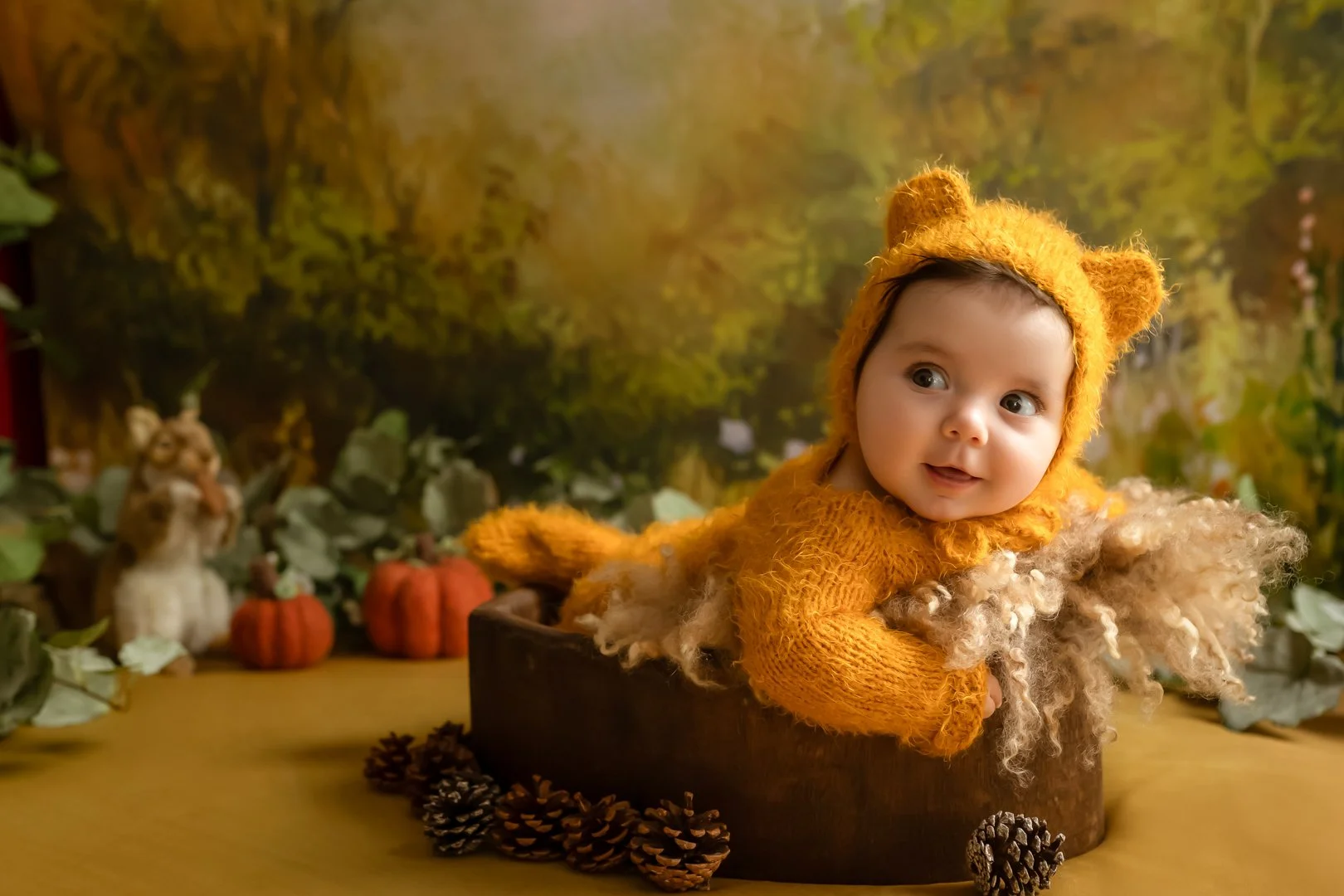 A baby dressed in a bear costume with a hood featuring bear ears, lying on a round platform surrounded by pinecones, with a fall-themed background including pumpkins, a dog plush, and autumn foliage.