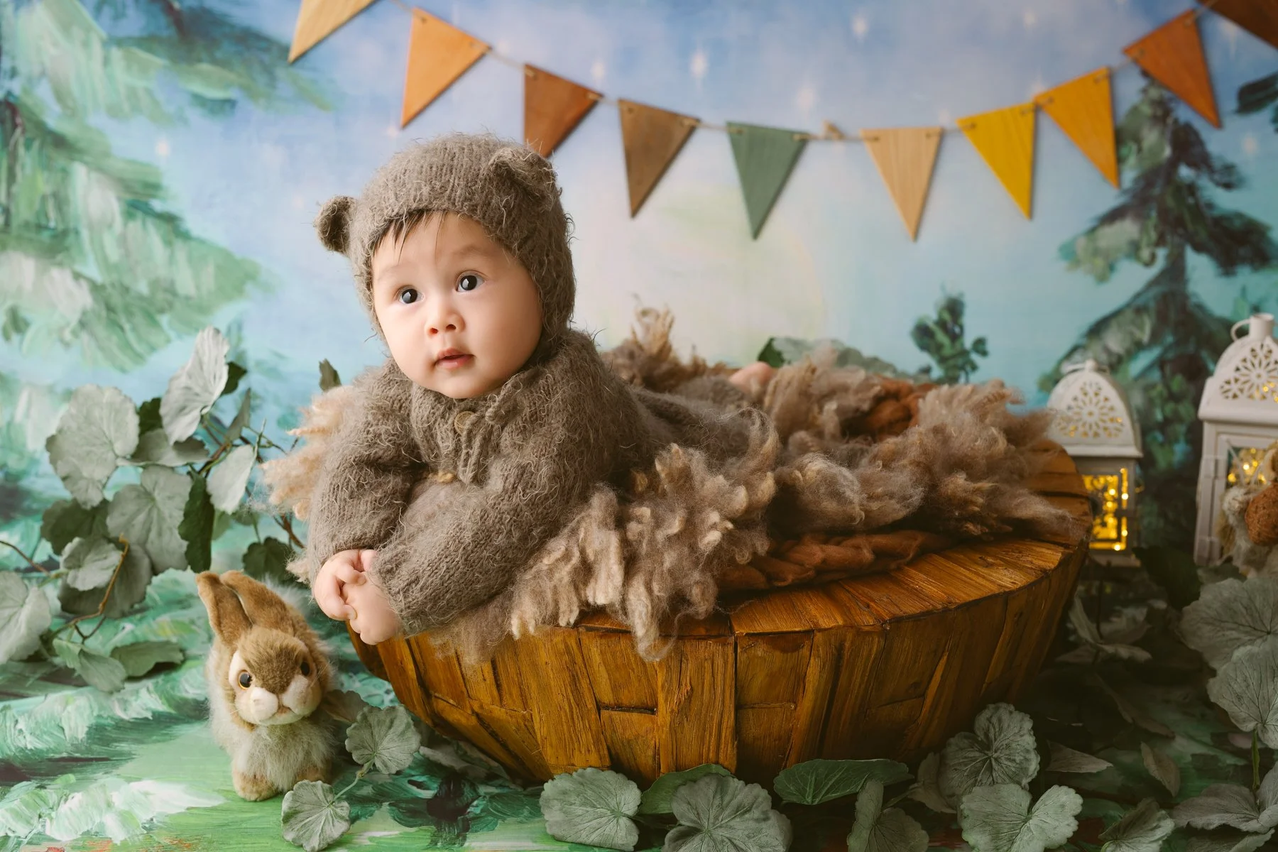 A toddler dressed in a brown, furry costume lying on a wooden boat, surrounded by a nature-themed backdrop with greenery and bunting flags, with a small plush rabbit toy nearby.