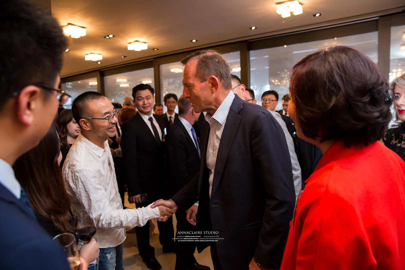 Man in dark suit shaking hands with a man in a white shirt at a professional networking event with people in suits and dresses in background.