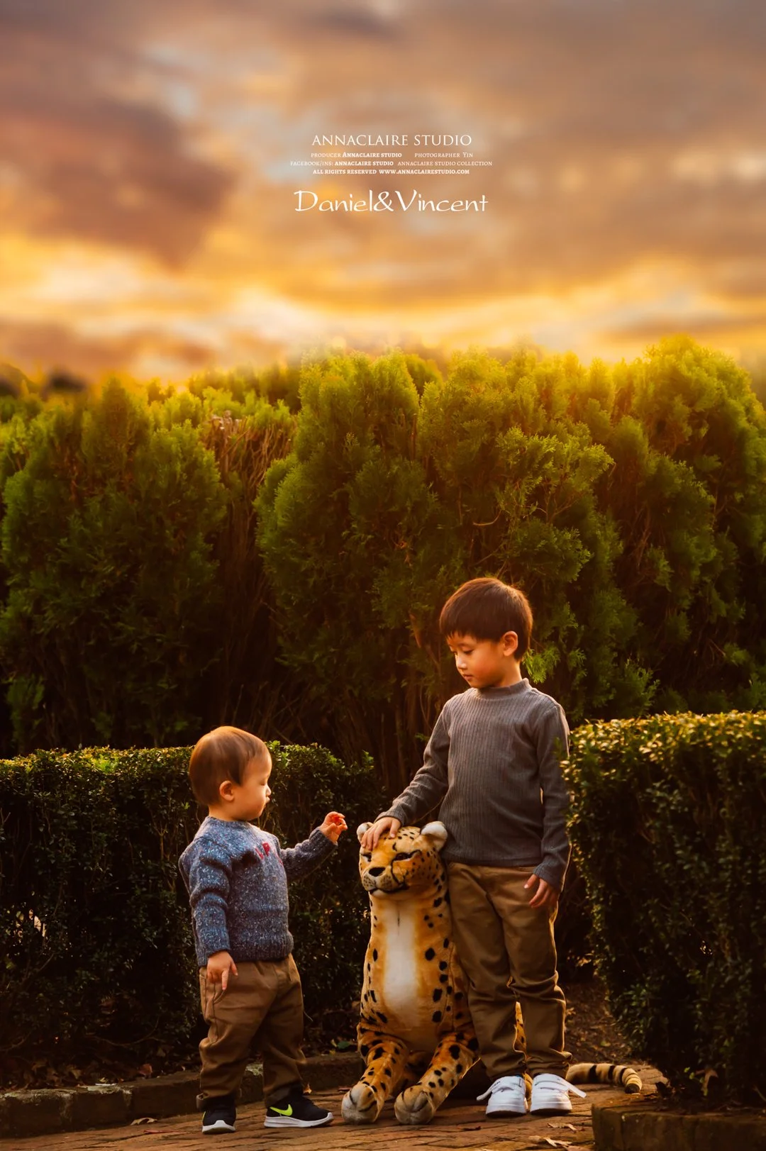 Two young boys, one younger and one older, standing outdoors next to a large plush toy jaguar. The older boy gently rests his hand on the plush toy while the younger boy reaches out towards it. They are surrounded by green bushes with a large bushy t