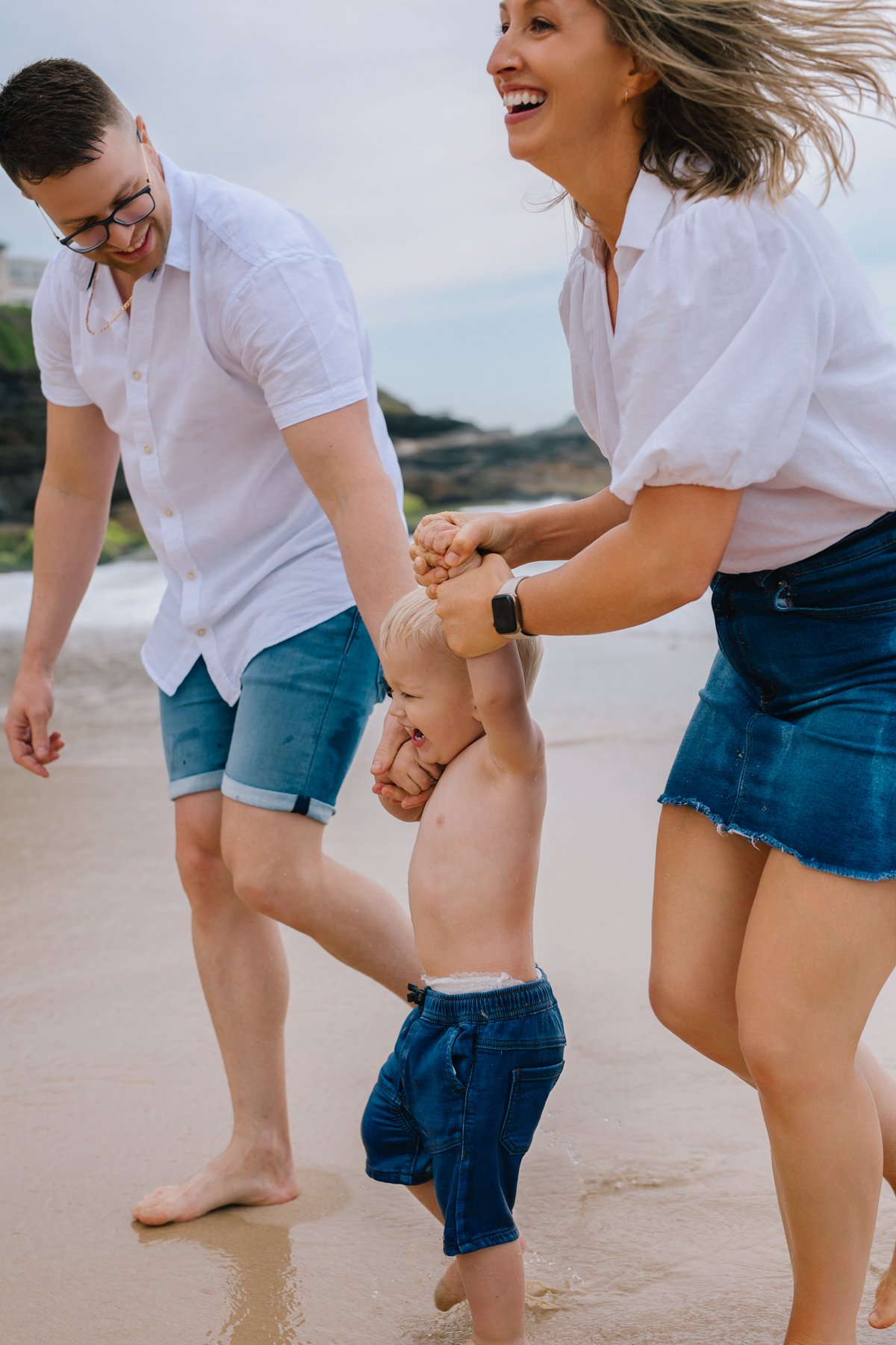 A family at the Bronte beach playing with young son, lifting him by the arms, all smiling and enjoying the moment.