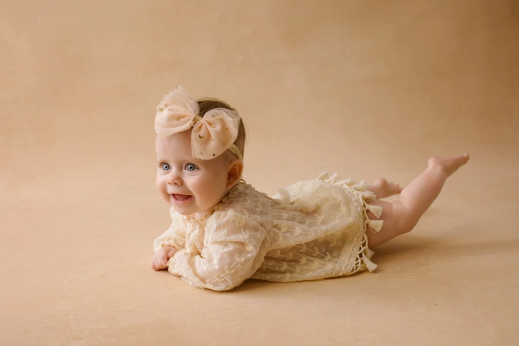A smiling baby girl with big blue eyes and light hair, wearing a cream-colored dress with lace and embroidery, and a large blush pink bow headband, lying on her stomach on a beige background.