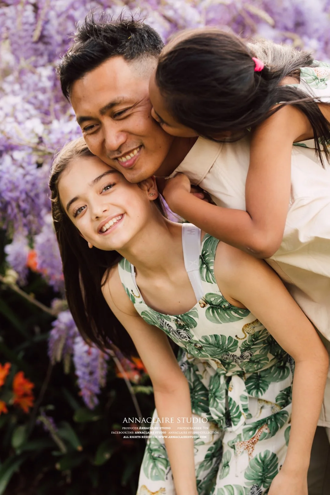 A man and two young girls sharing a joyful moment outdoors with purple flowers in the background.