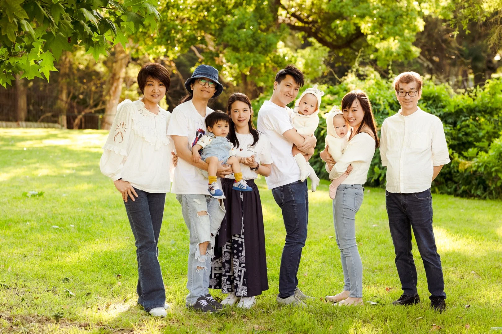 Family of eight people, including children, standing outdoors in a green park with trees in the background.