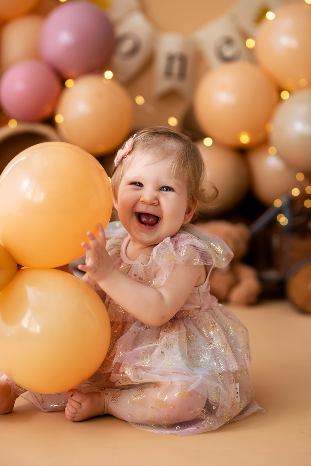 A happy baby girl sitting on the floor, holding orange balloons, with a cake smash photo background decorated with pink and beige balloons and string lights.