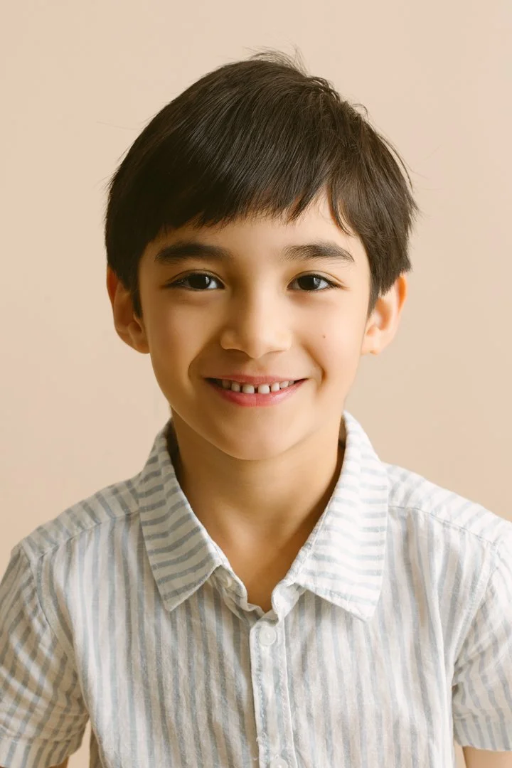 Portrait of a young boy with dark hair wearing a light-colored, striped shirt, smiling against a plain beige background.