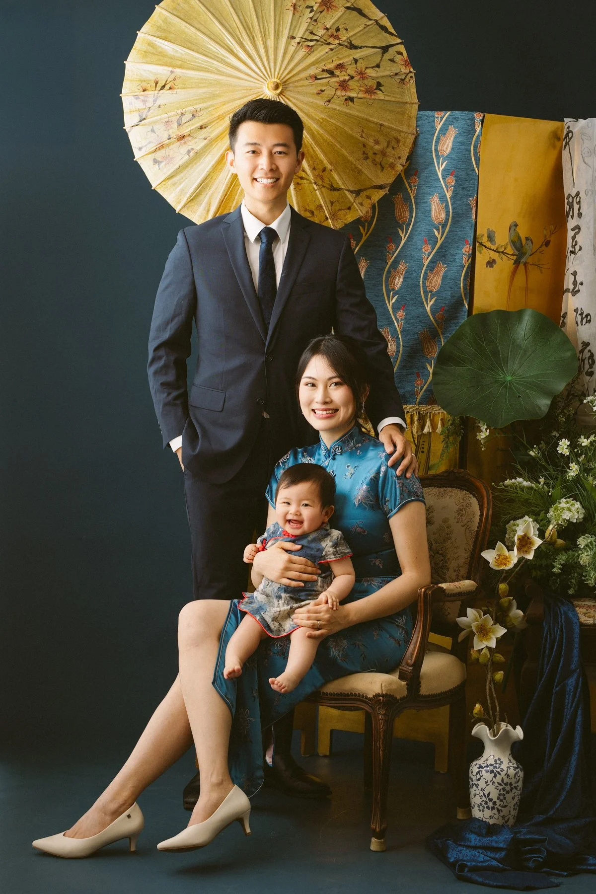 A family portrait with a man in a suit, a woman in a traditional blue dress, and a smiling baby. The woman is seated on a vintage chair, holding the baby, and the man stands behind her with his hand on her shoulder. The background features Asian-them