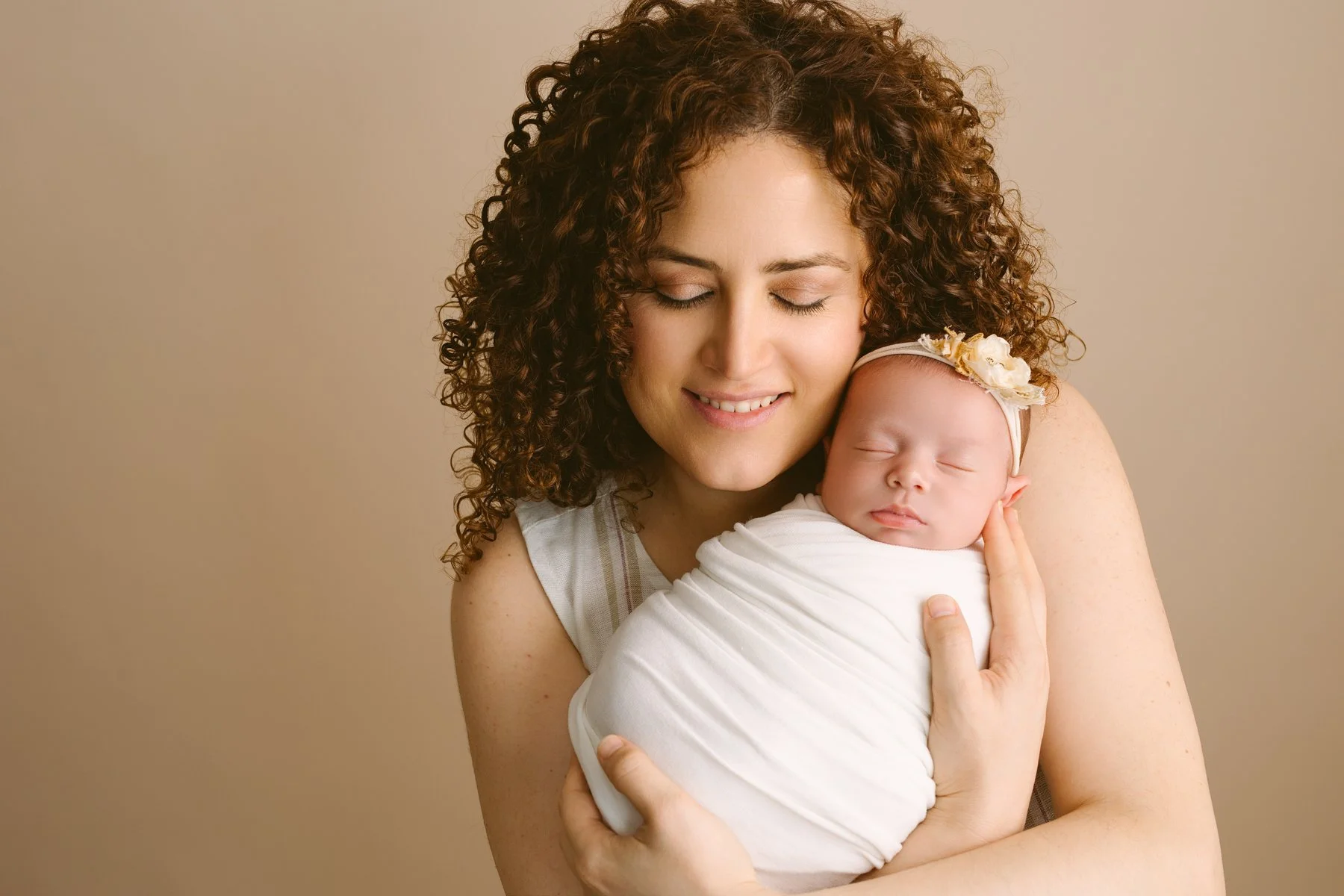 A woman with curly hair holding a sleeping baby wrapped in a white blanket, both smiling gently with eyes closed.