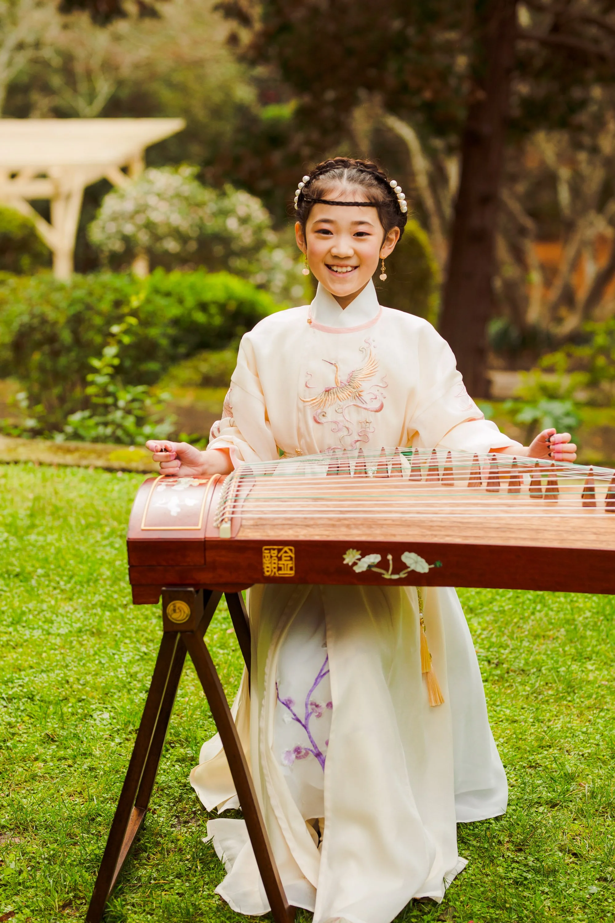 Young girl wearing traditional chinese outfits= playing a guzheng outdoors in a garden.