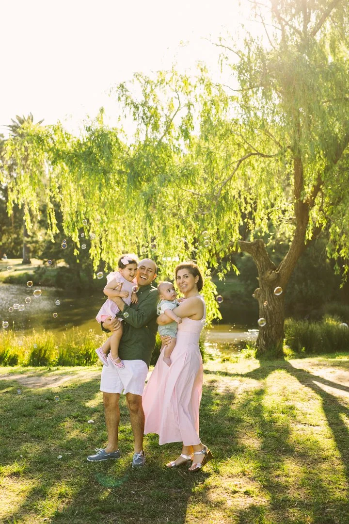 A happy family of four standing outdoors in a park during the daytime. The father is holding a young girl, while the mother is holding a baby. They are smiling and enjoying the sunny weather with a large green tree and a pond in the background.