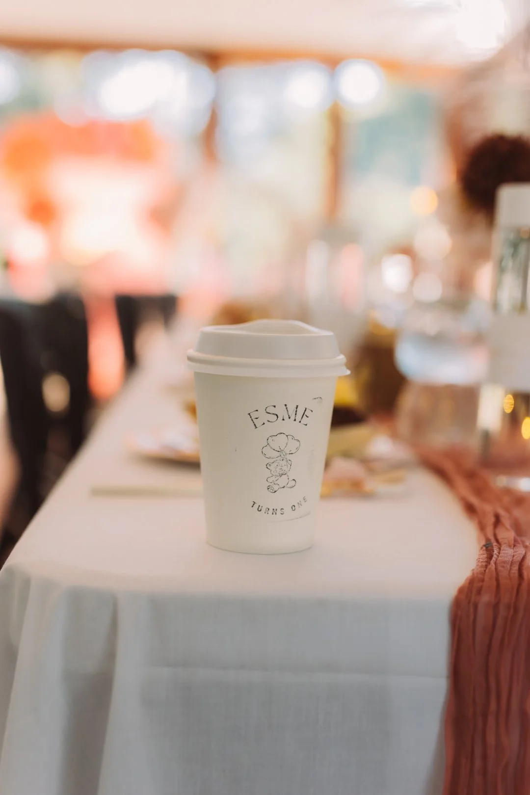 A white disposable coffee cup with the name "ESME" and a cartoon character drawing, placed on a table at a celebration or gathering.