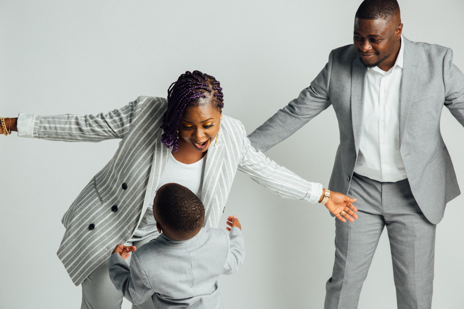 A happy family of three, with a woman, man, and young boy, playing together and smiling. The woman has braided purple hair and is wearing a grey pinstripe blazer, the man is in a light grey suit, and the boy is in a light grey suit, all against a pla