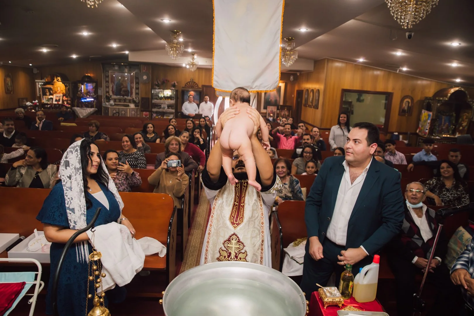 A priest holding a naked baby in a church during a religious ceremony, while the congregation, dressed in formal and semi-formal clothing, watches and takes photos.