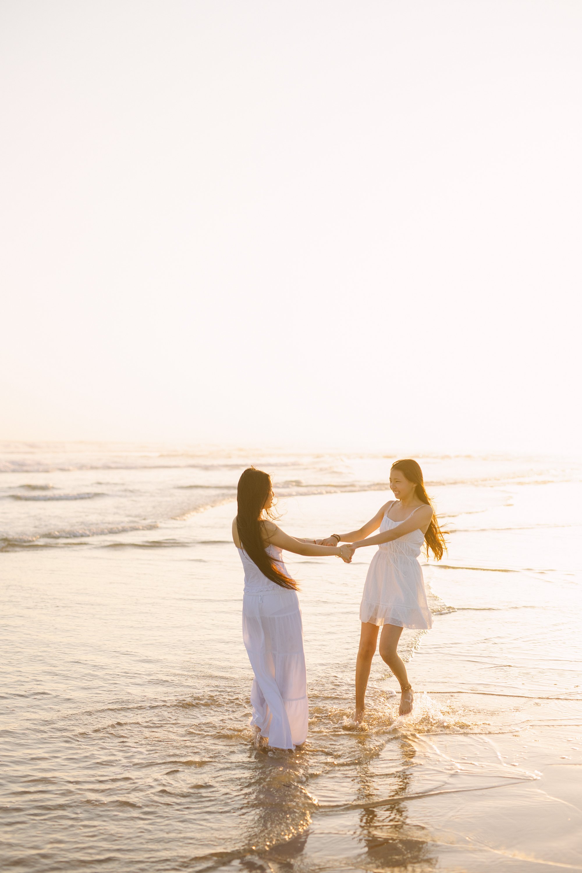 Two women in white dresses holding hands and playing in the ocean at sunset.