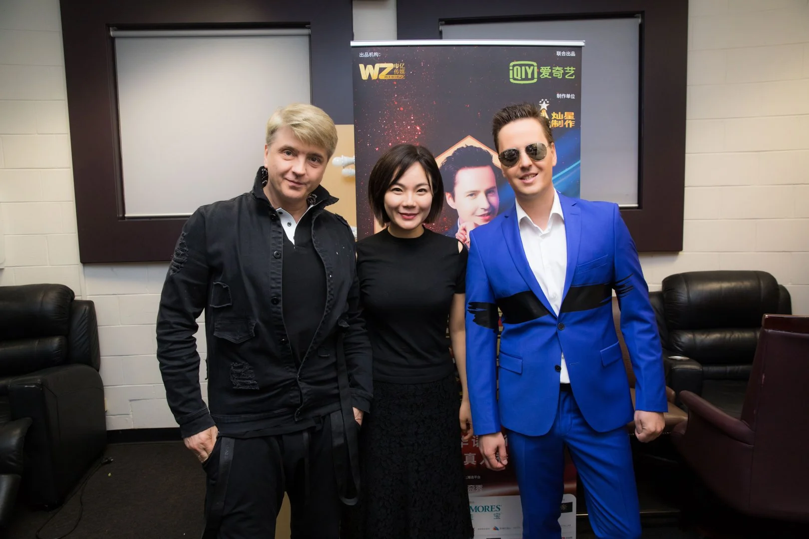 Three people standing together indoors, posing for a photo in front of a promotional poster with Chinese characters. The person on the left is a man with blond hair wearing a black jacket and black pants. The person in the middle is an Asian woman wi