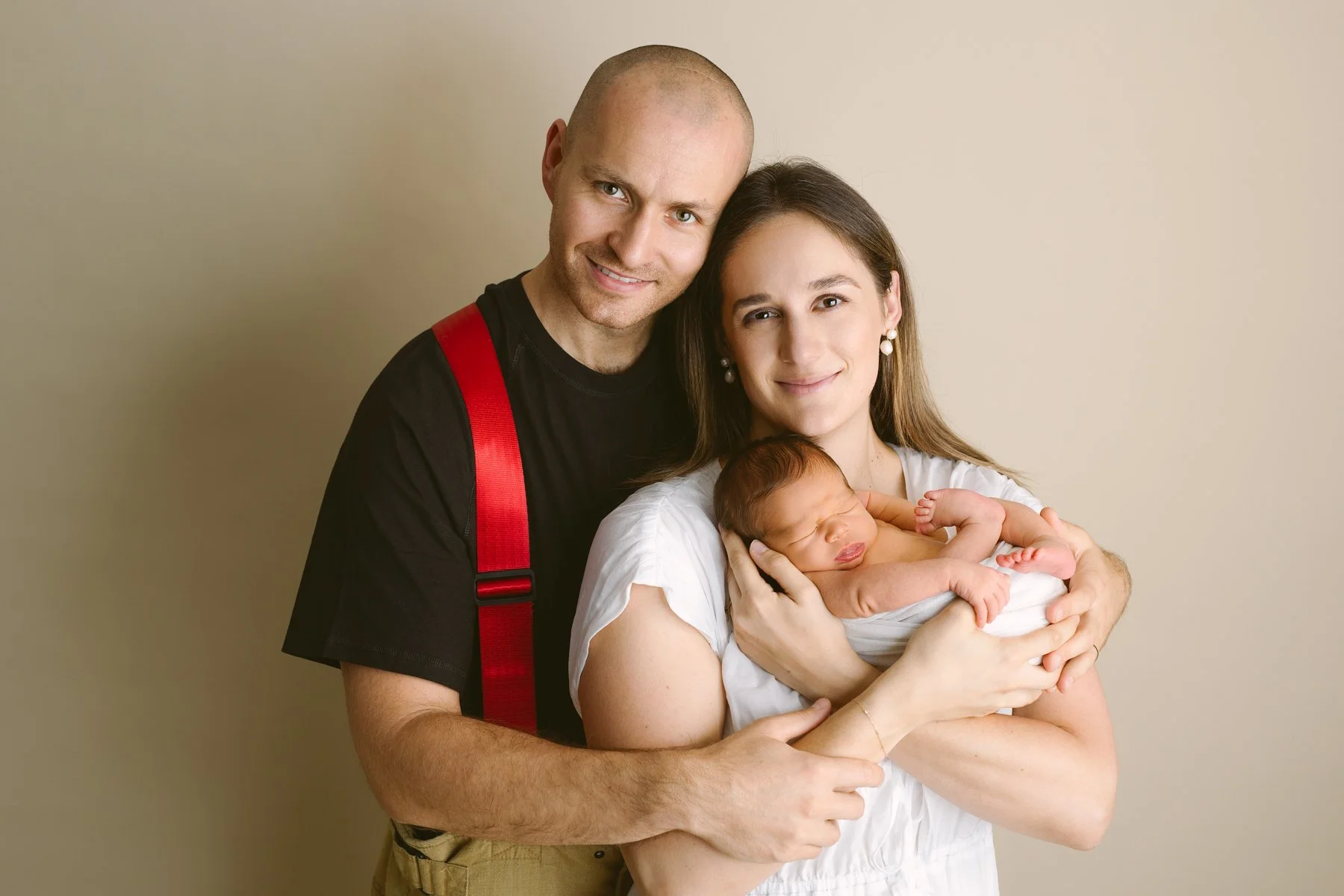 A family of three, including mum holding a newborn baby boy, with dad standing behind them with love, smiling against a plain beige background.