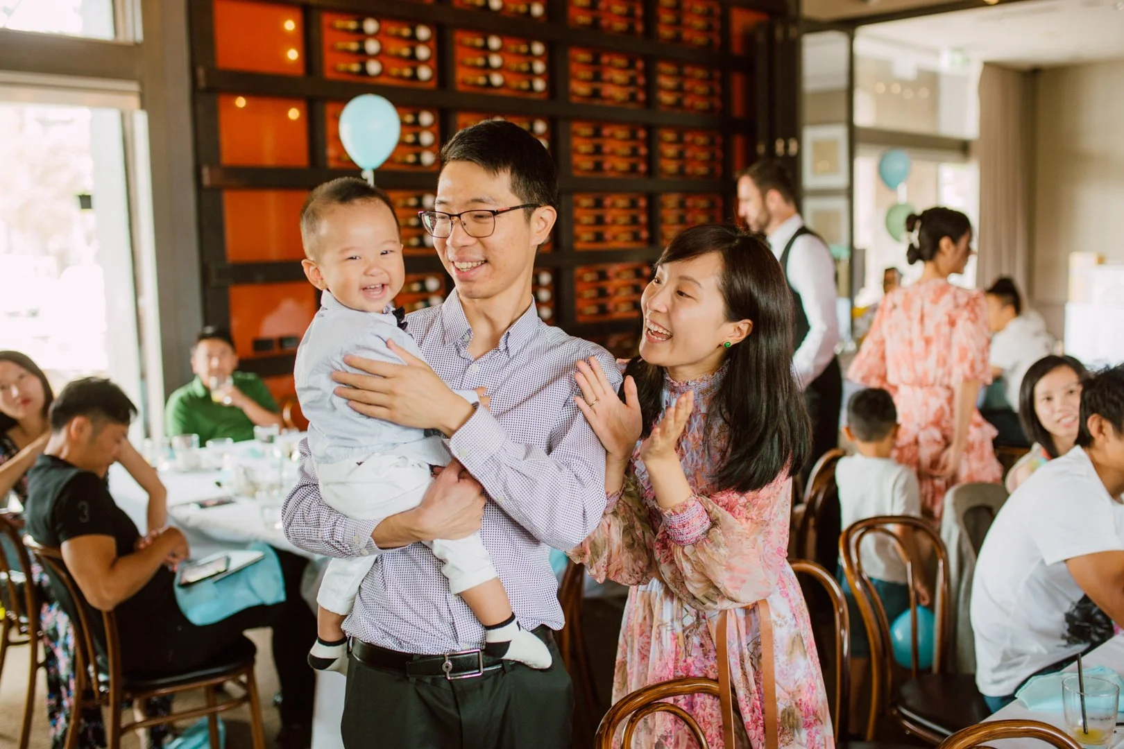 A young Asian man holding a smiling toddler at a lively celebration, with a woman beside him smiling and touching his shoulder, in a restaurant decorated with balloons and a wine rack in the background, other guests sitting and talking at tables.